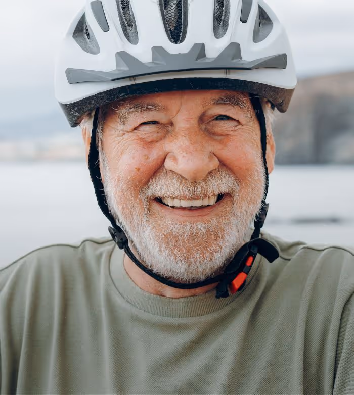 Smiling elderly man wearing a white bike helmet and green shirt outdoors near water.