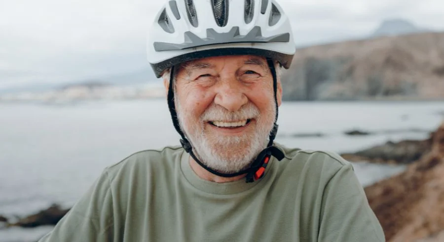 Smiling elderly man wearing a white bike helmet outdoors near a rocky shoreline and water.
