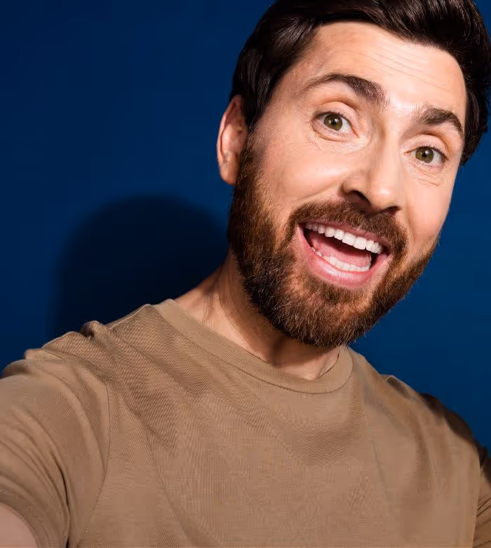 Smiling bearded man wearing a tan shirt posing against a dark blue background.