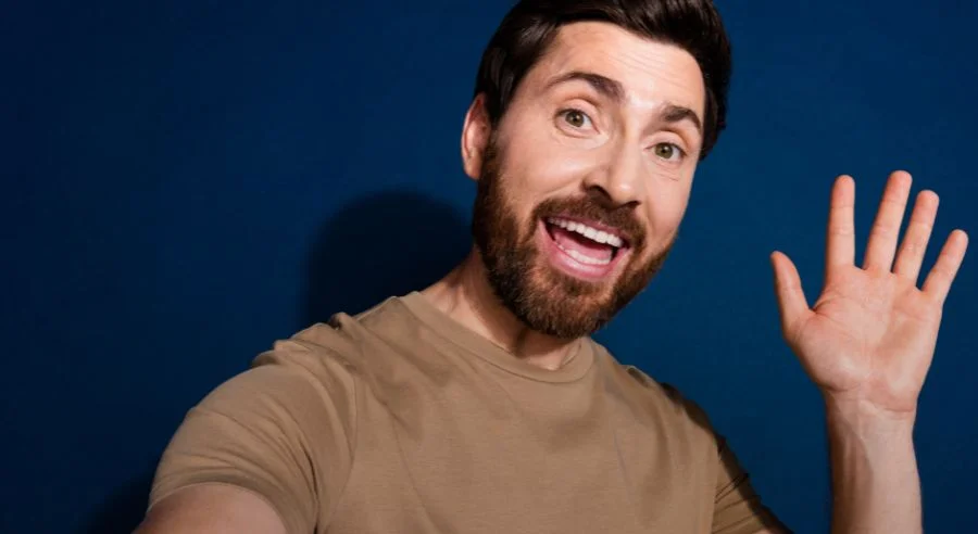 Bearded man smiling and waving with open hand against a dark blue background.