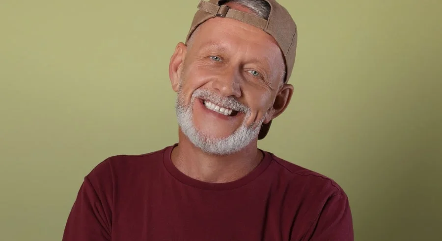 Smiling older man with a gray beard wearing a backwards beige cap and maroon shirt against a light green background.