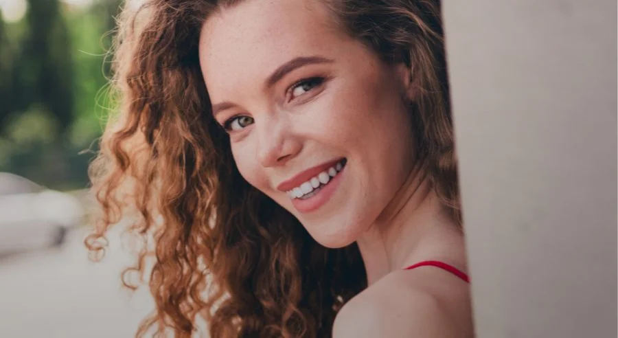 Smiling young woman with curly hair leaning against a wall in an outdoor setting.