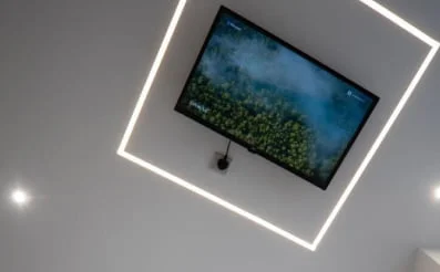 Ceiling-mounted TV screen with a forest image, framed by square LED light fixture on white ceiling.