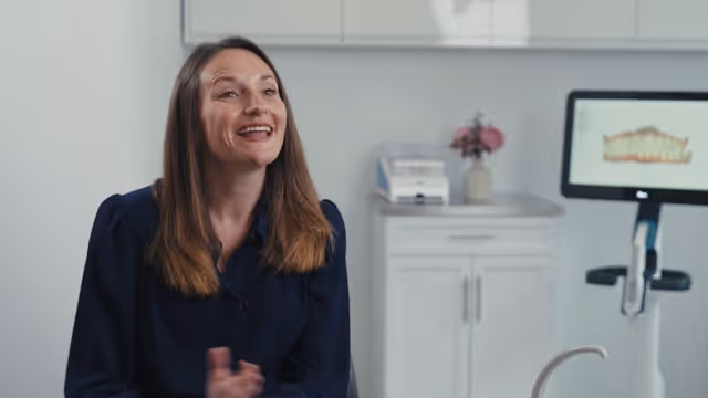 Woman with long brown hair smiling in a modern dental office with a dental model on screen behind her.