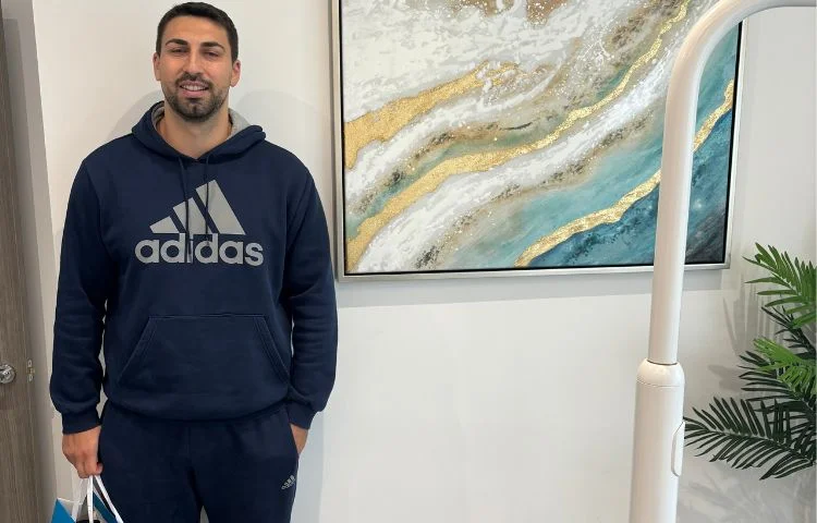 Man in navy Adidas hoodie and pants stands smiling beside abstract wall art and a potted plant.