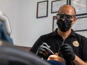 Dentist wearing black mask and gloves treating a patient in a dental clinic with certificates on the wall.