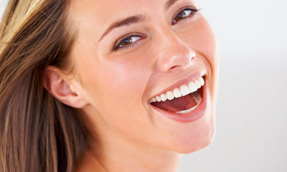 Close-up of a smiling woman with brown hair and bright white teeth against a light background.