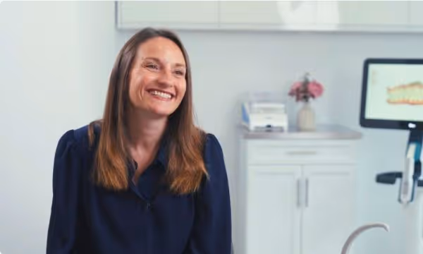 Smiling woman with long brown hair sitting in a bright dental office.
