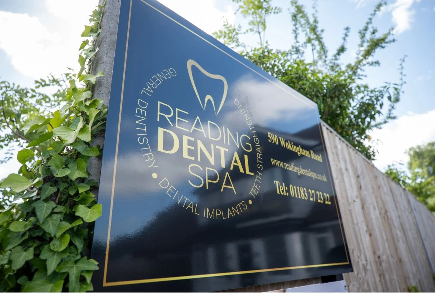 Outdoor sign for Reading Dental Spa with contact info and location on a fence with greenery.