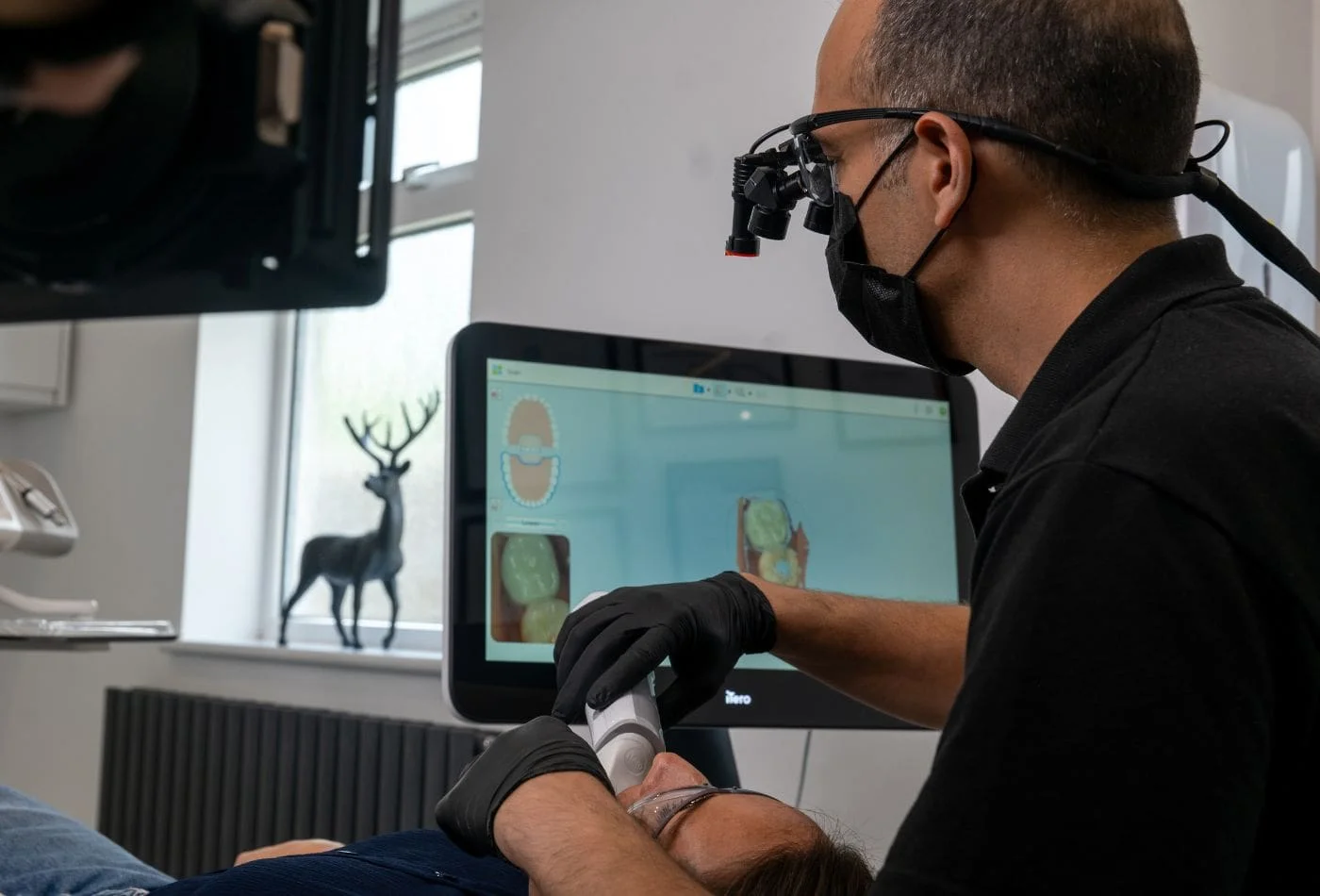 Dentist wearing magnifier glasses and mask scans patient's teeth using dental intraoral scanner.