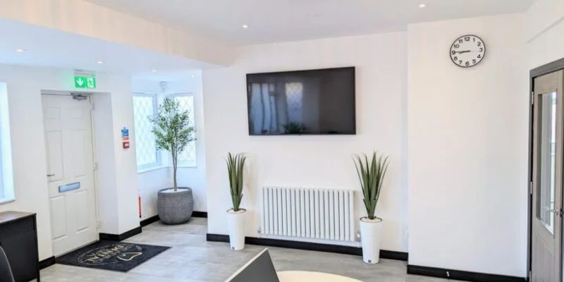 Minimalist waiting area with wall clock, flat screen TV, two tall potted plants, and white door with exit sign.