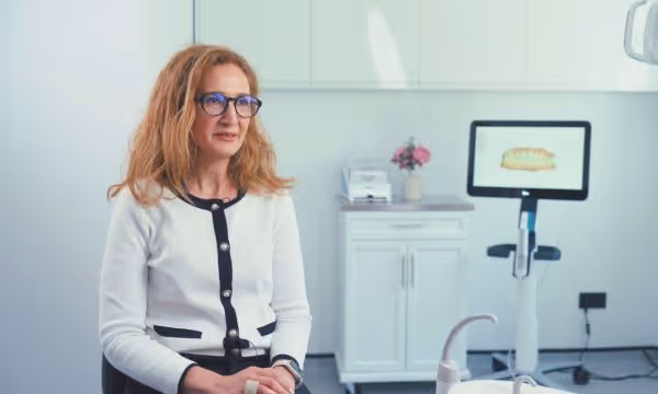 Woman with glasses sitting in a dental office with digital dental scan equipment in the background.