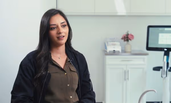 Woman with long dark hair speaking while seated in a modern dental office with equipment behind her.