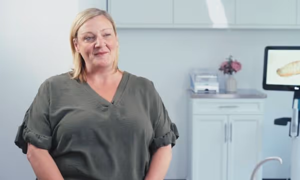 Smiling woman with short blonde hair wearing a gray blouse in a bright room with white cabinets.