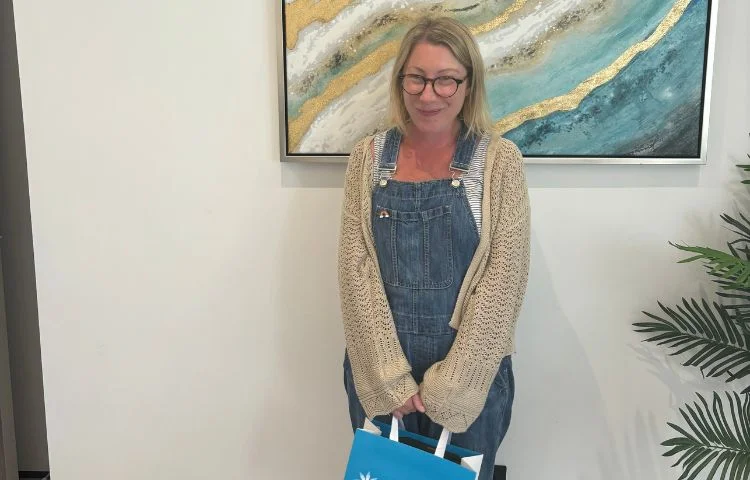 Smiling woman with glasses in denim overalls and beige sweater holding a blue and white shopping bag.