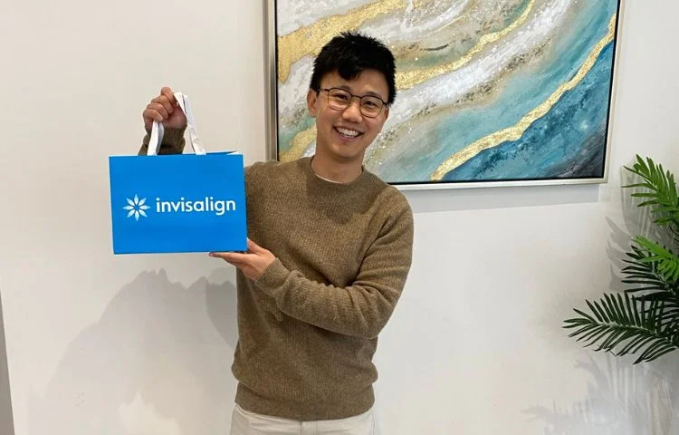 Smiling man holding a blue Invisalign shopping bag in a room with artwork and a plant.