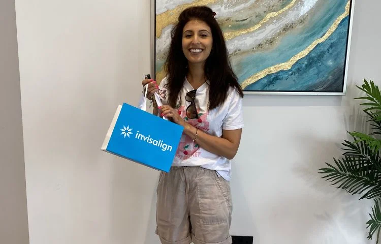 Smiling woman holding a blue Invisalign bag standing indoors by a plant and wall art.