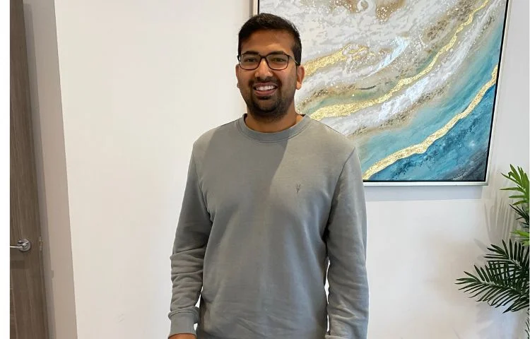 Man with glasses smiling, wearing a gray sweatshirt, standing indoors near abstract art and a plant.