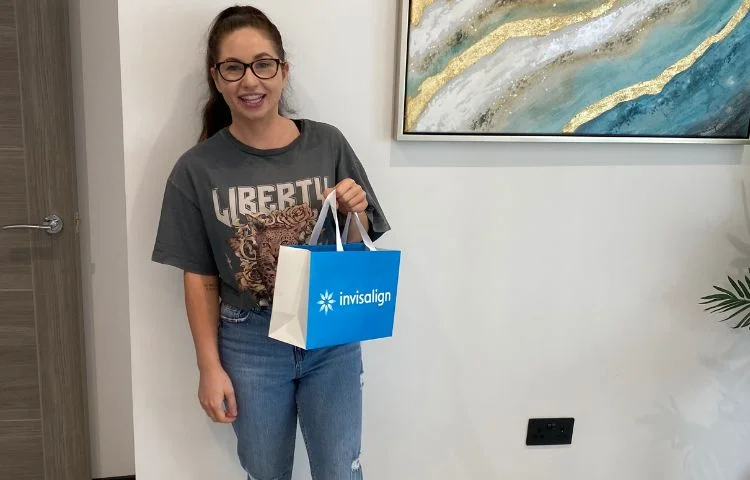 Smiling woman in glasses holding an Invisalign branded shopping bag, standing indoors near a door.