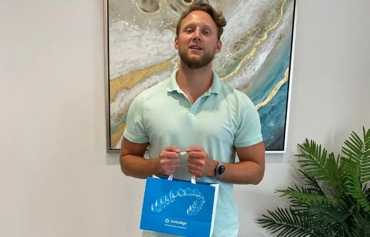 Man in light blue polo shirt holding a blue Invisalign bag with a decorative painting and plant behind him.