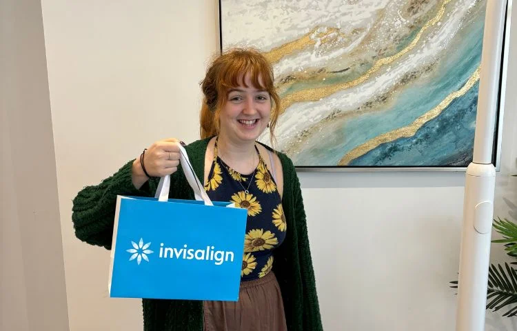 Smiling woman holding a blue Invisalign gift bag indoors next to abstract wall art.