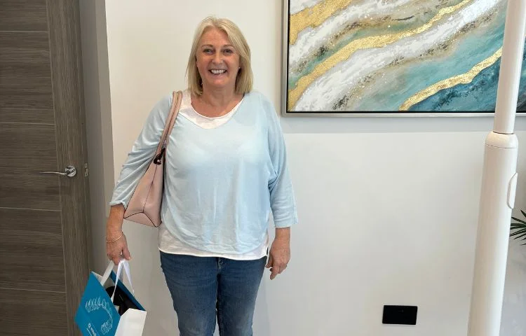 Smiling woman with blond hair holding shopping bags and a pink purse standing indoors next to a painting.