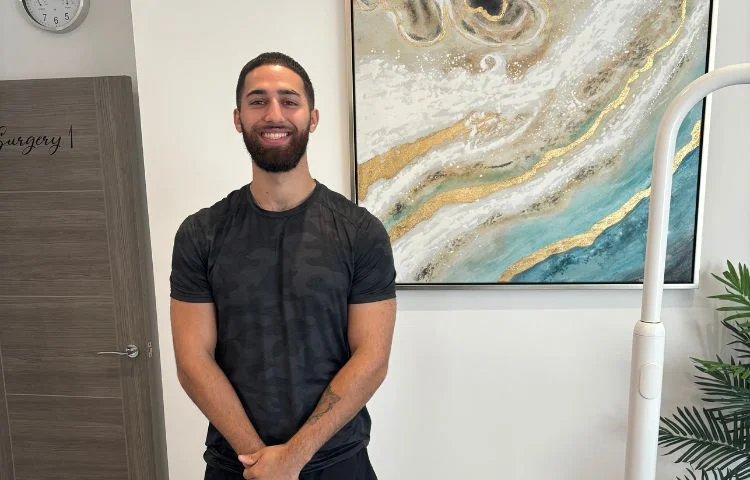 Smiling man in black t-shirt standing in a room with abstract wall art and a wooden door labeled Surgery.
