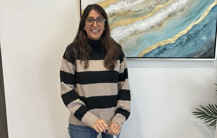 Smiling woman with glasses in black and beige striped sweater standing indoors near a blue and gold painting.