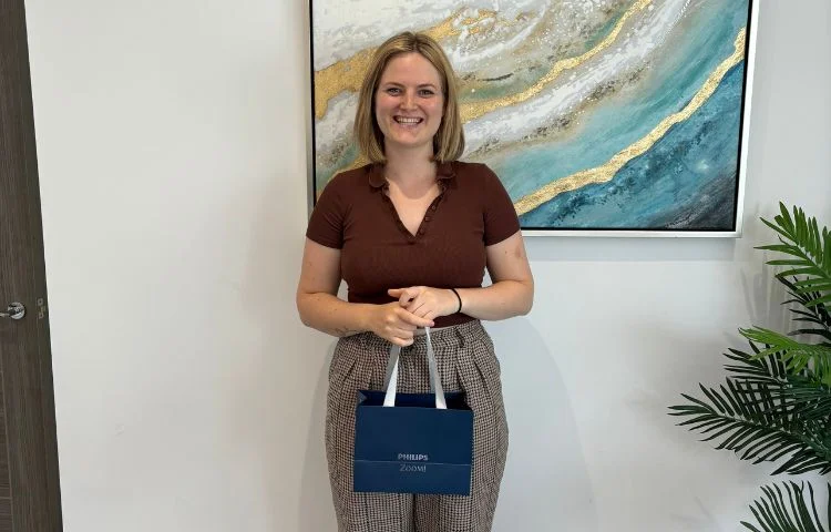 Smiling woman standing indoors holding a dark blue Philips Zoom bag with an abstract painting behind her.