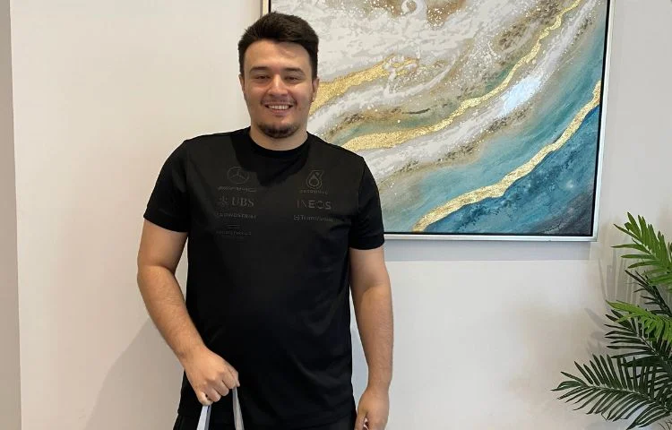 Smiling man in black t-shirt standing indoors near blue and gold abstract wall art and green plant.