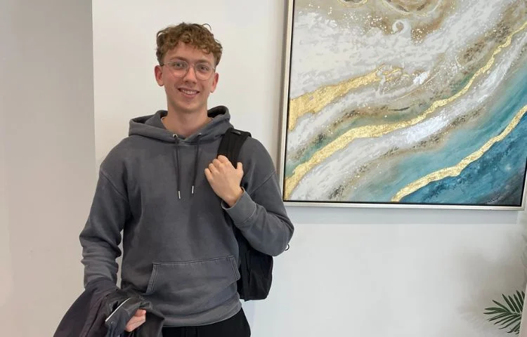 Young man wearing glasses and a gray hoodie, smiling and holding a jacket and backpack strap indoors.