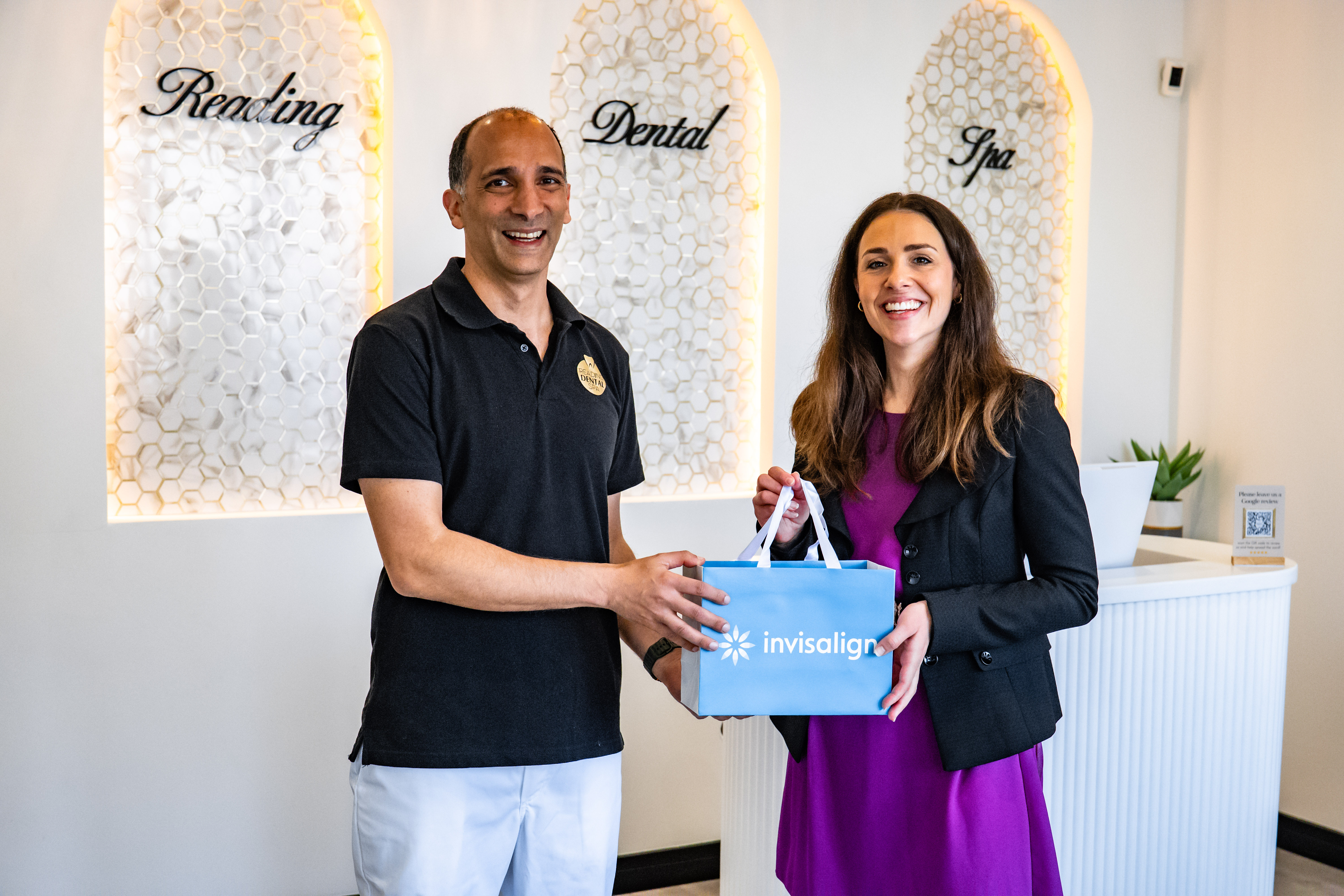 Man in dental polo handing a blue Invisalign bag to a smiling woman in purple dress and black blazer.
