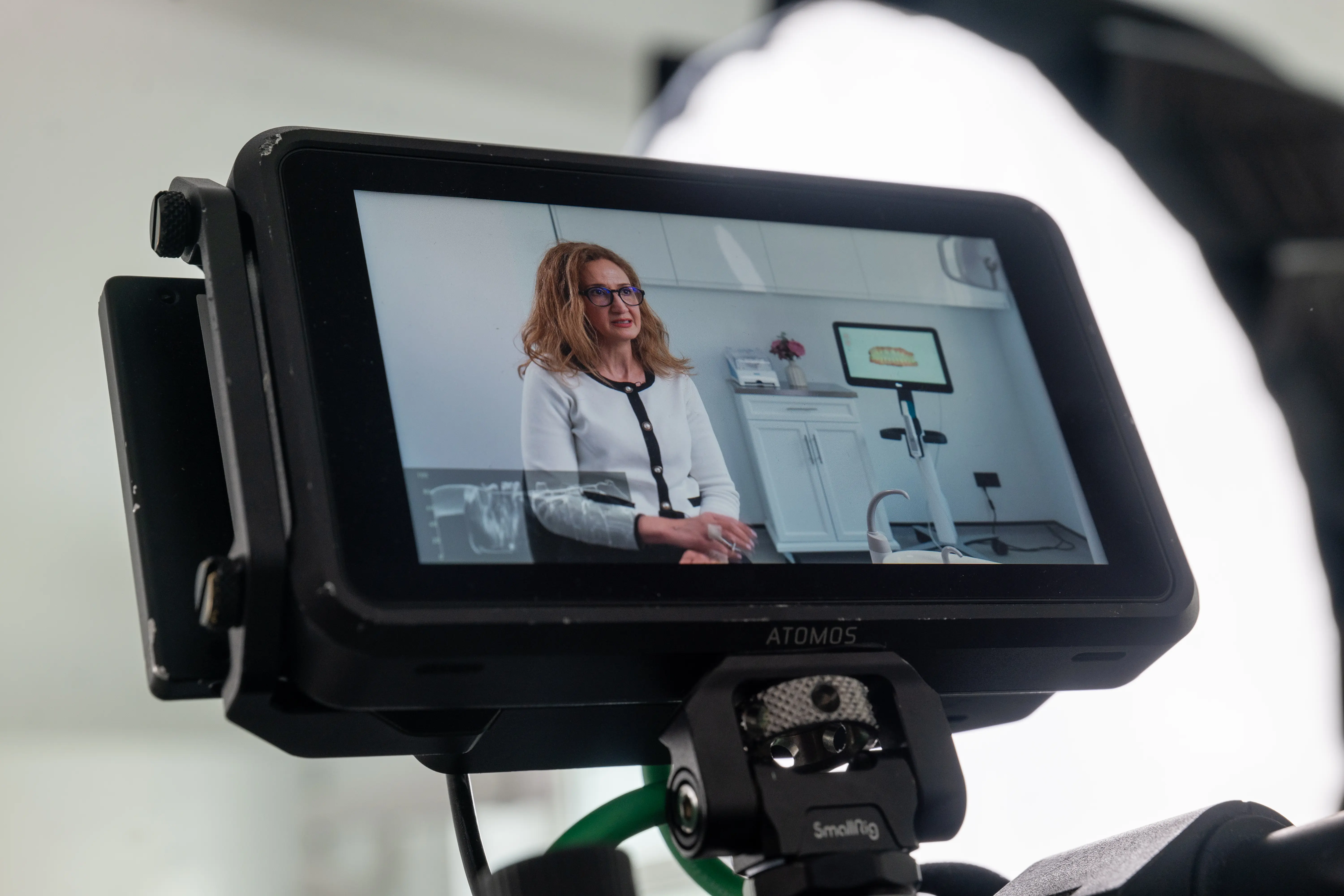 Video camera monitor showing a woman with glasses in a white and black jacket sitting in a dental clinic.