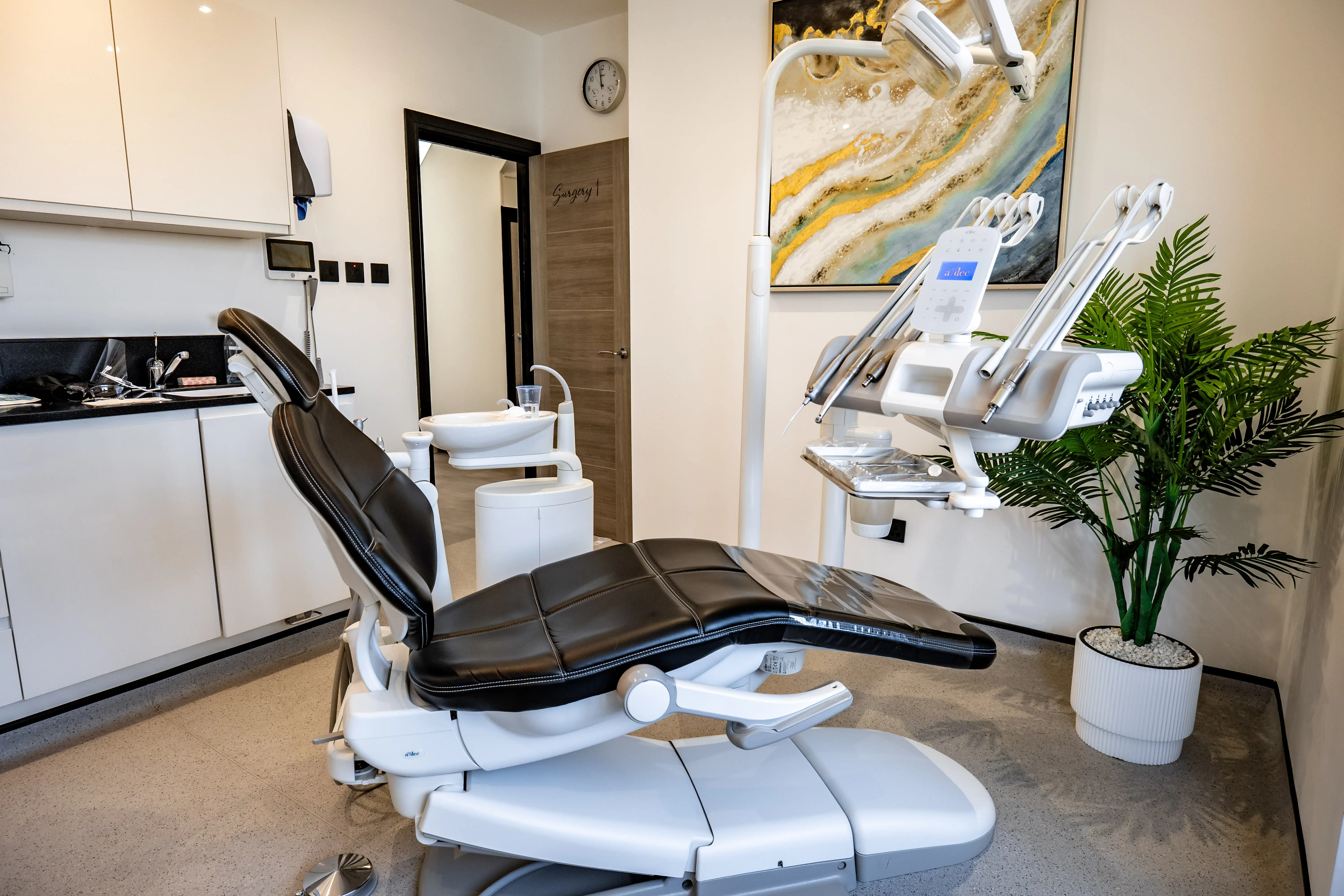 Modern dental treatment room with black dental chair, equipment, sink, and potted plant.