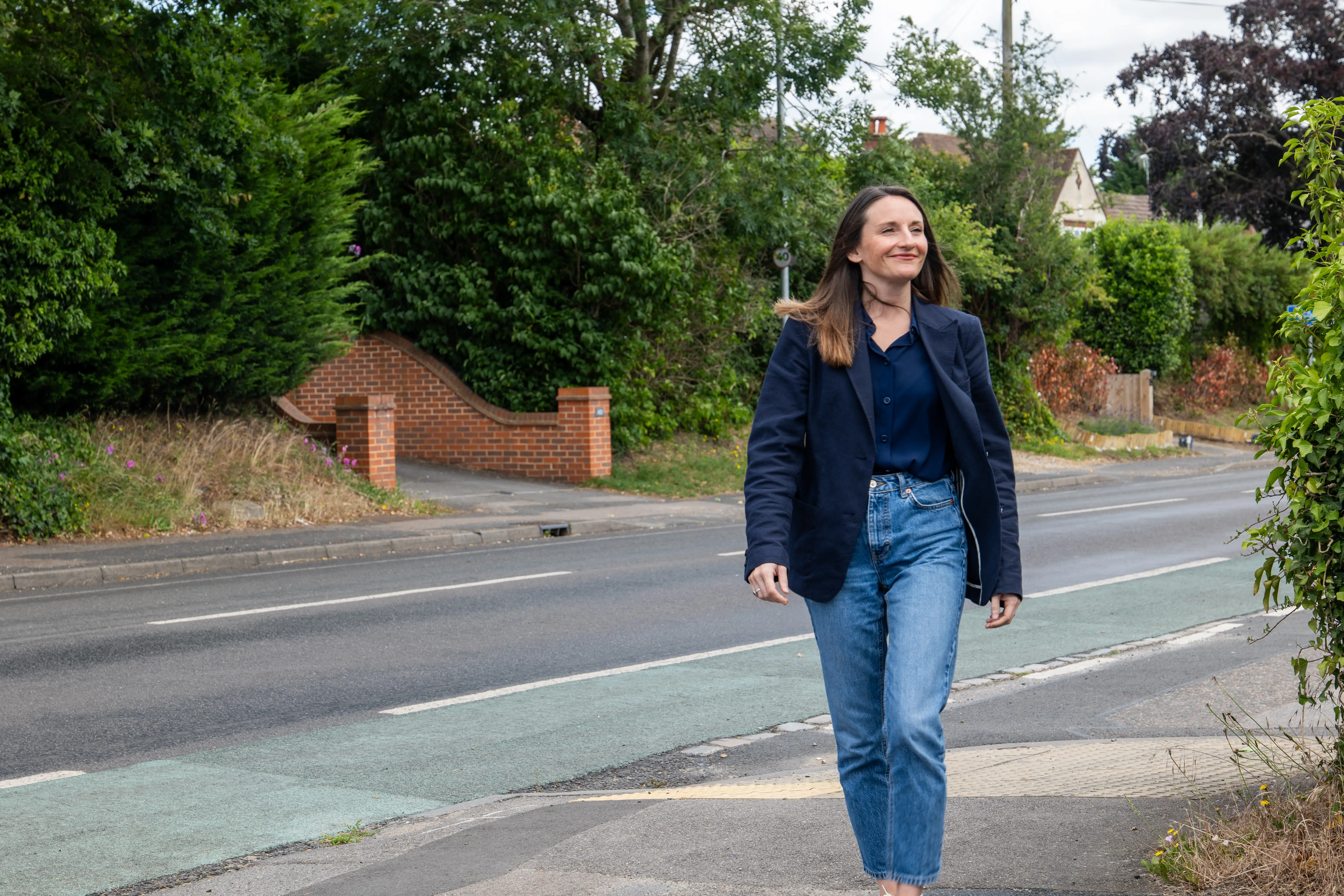 Woman in a navy blazer and jeans walking on a sidewalk next to a road with greenery.