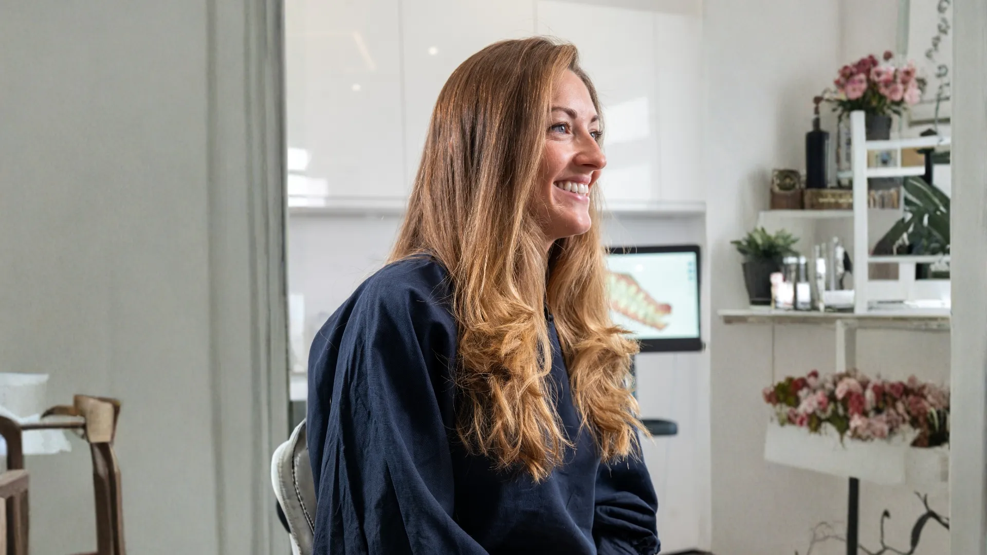 Smiling woman with long wavy hair wearing a dark shirt seated indoors near flowers and a computer screen.