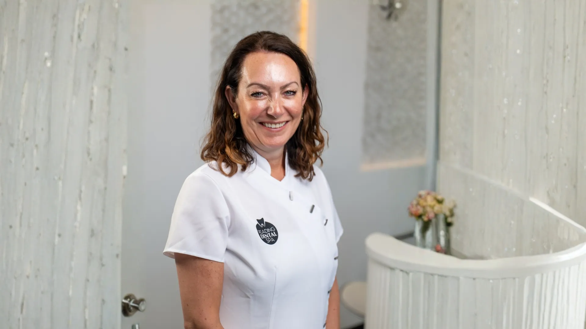 Smiling woman in white uniform with Reading Dental Spa logo standing in a spa reception area.