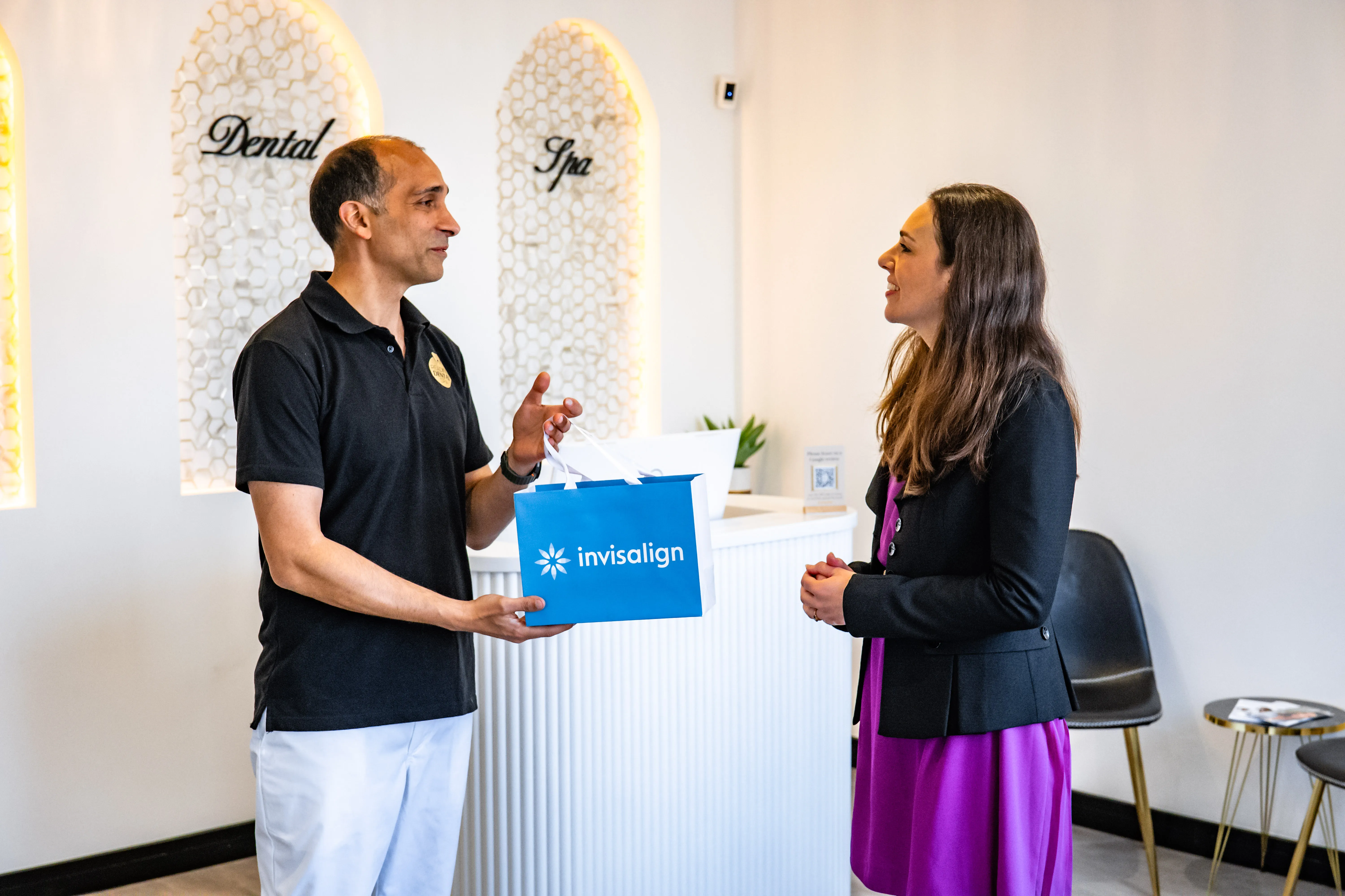 Man in dental spa black polo handing woman in purple dress a blue Invisalign bag at reception.