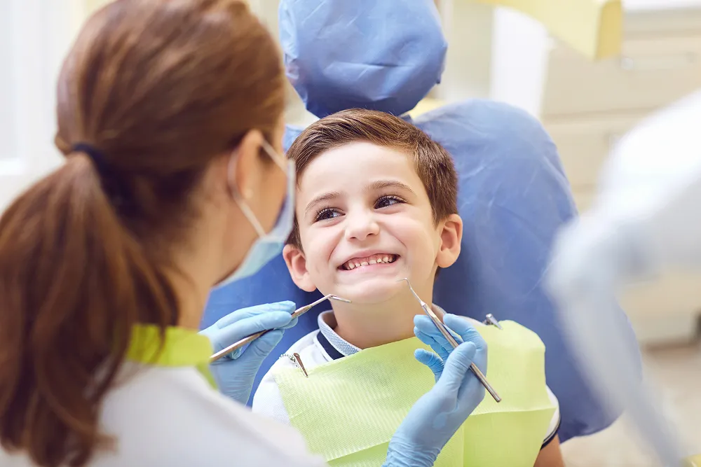 Smiling young boy sitting in a dental chair with a dentist holding dental tools near his mouth.