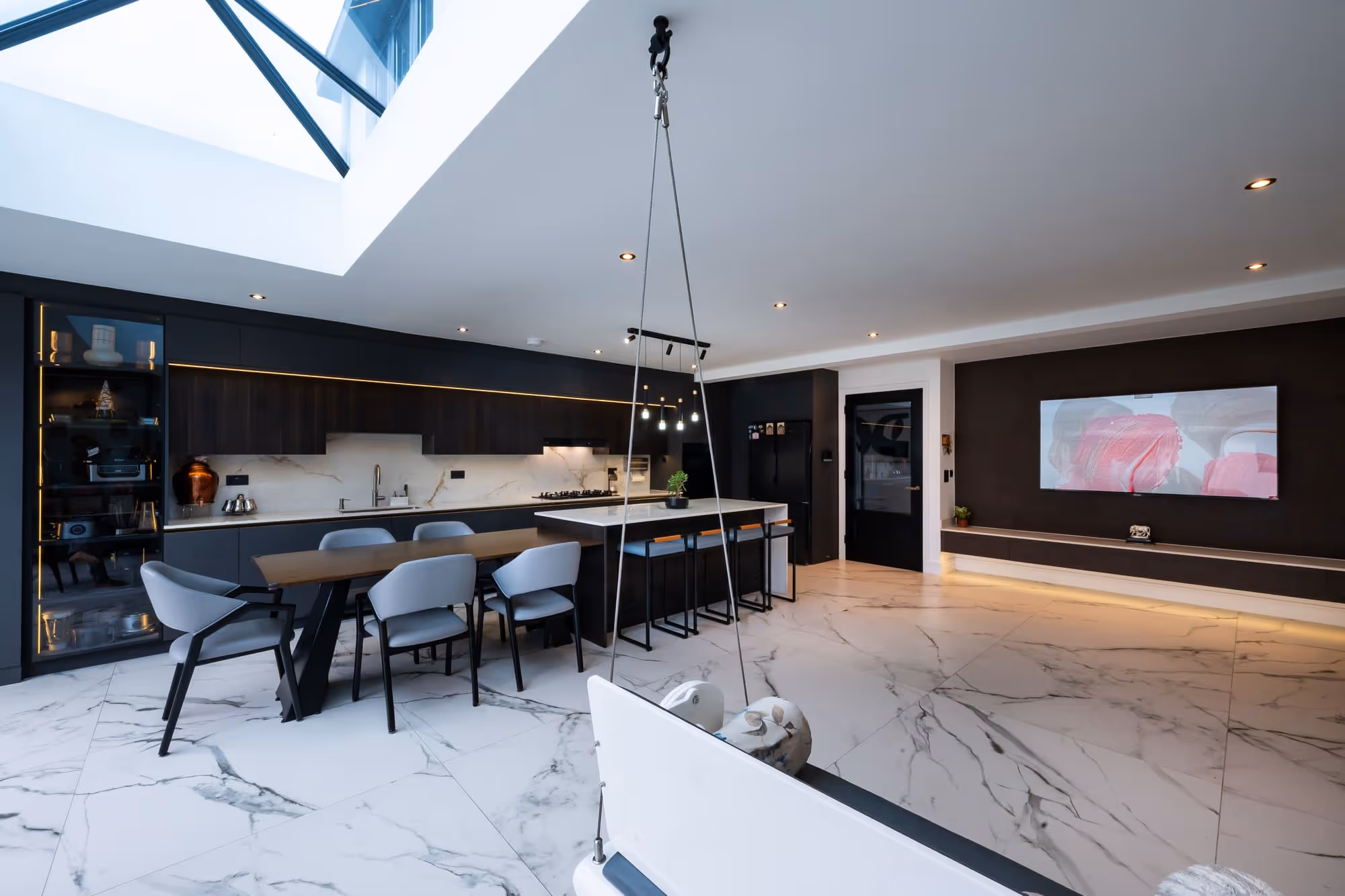 Modern kitchen and dining area with black cabinets, marble floor, and hanging lights under skylight.
