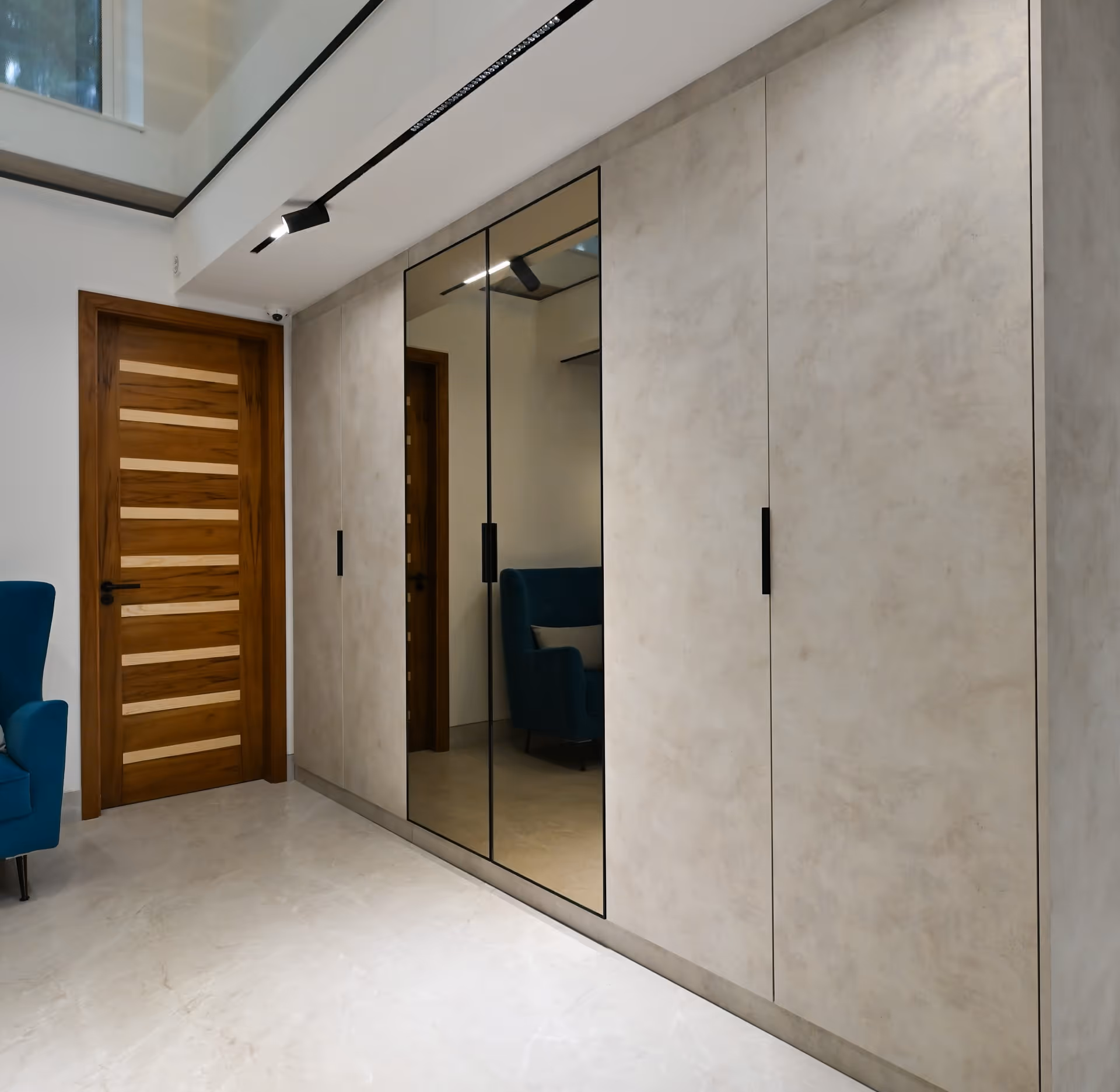 Modern hallway with wooden door, beige wall cabinets, and a blue armchair reflected in mirrored doors.