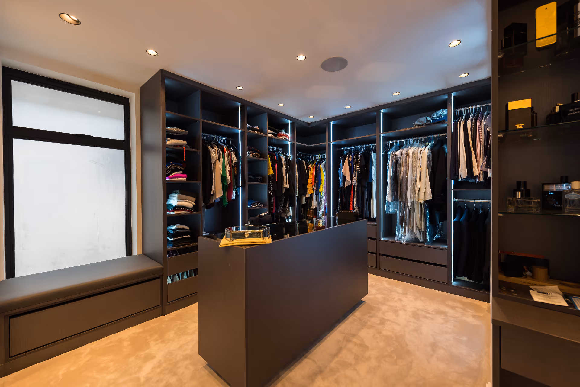 Modern walk-in closet with dark shelves, stacked clothes, hanging garments, and a central island.