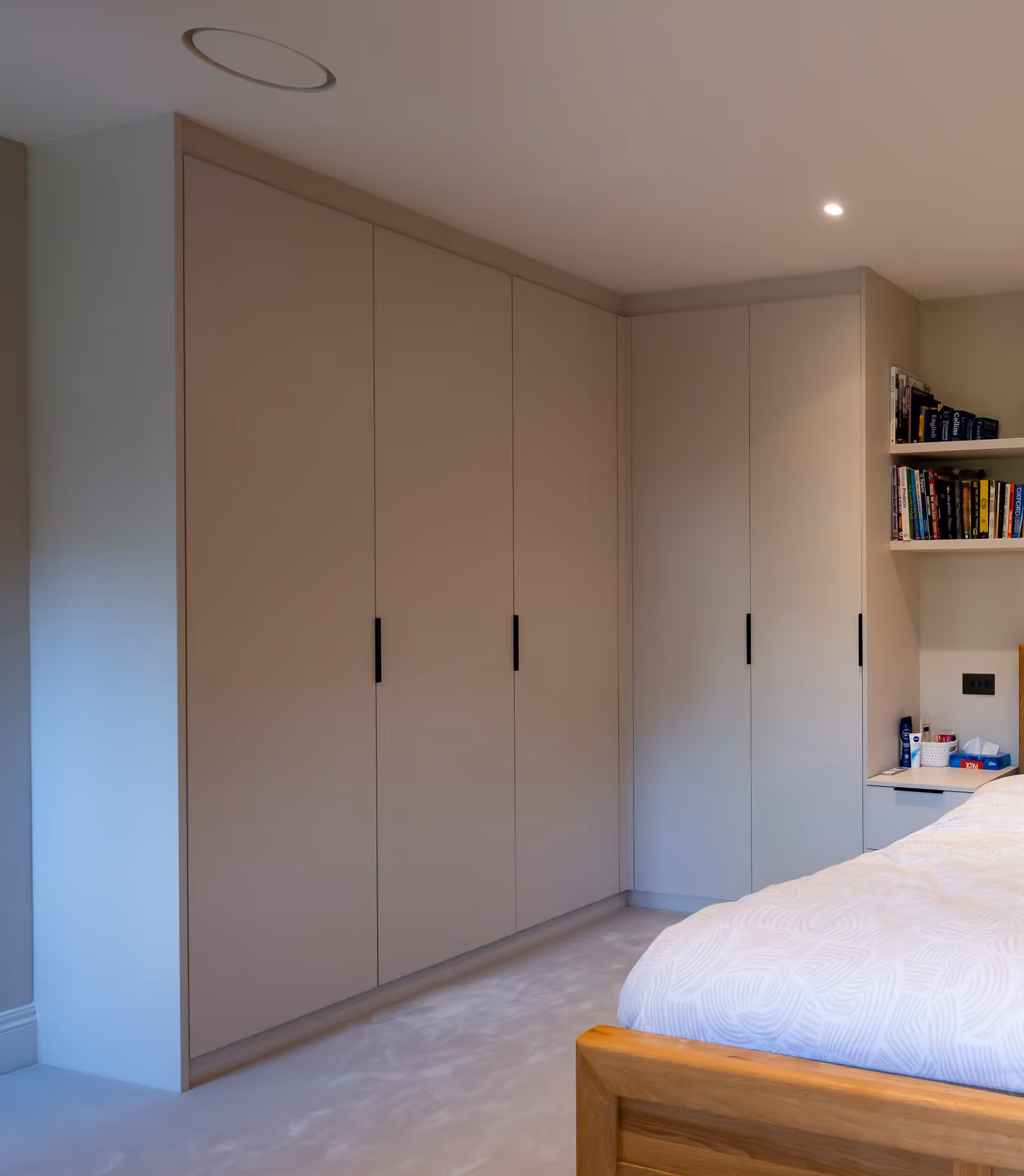 Corner bedroom with built-in beige wardrobes, bookshelf, and a wooden bedframe with white bedding.