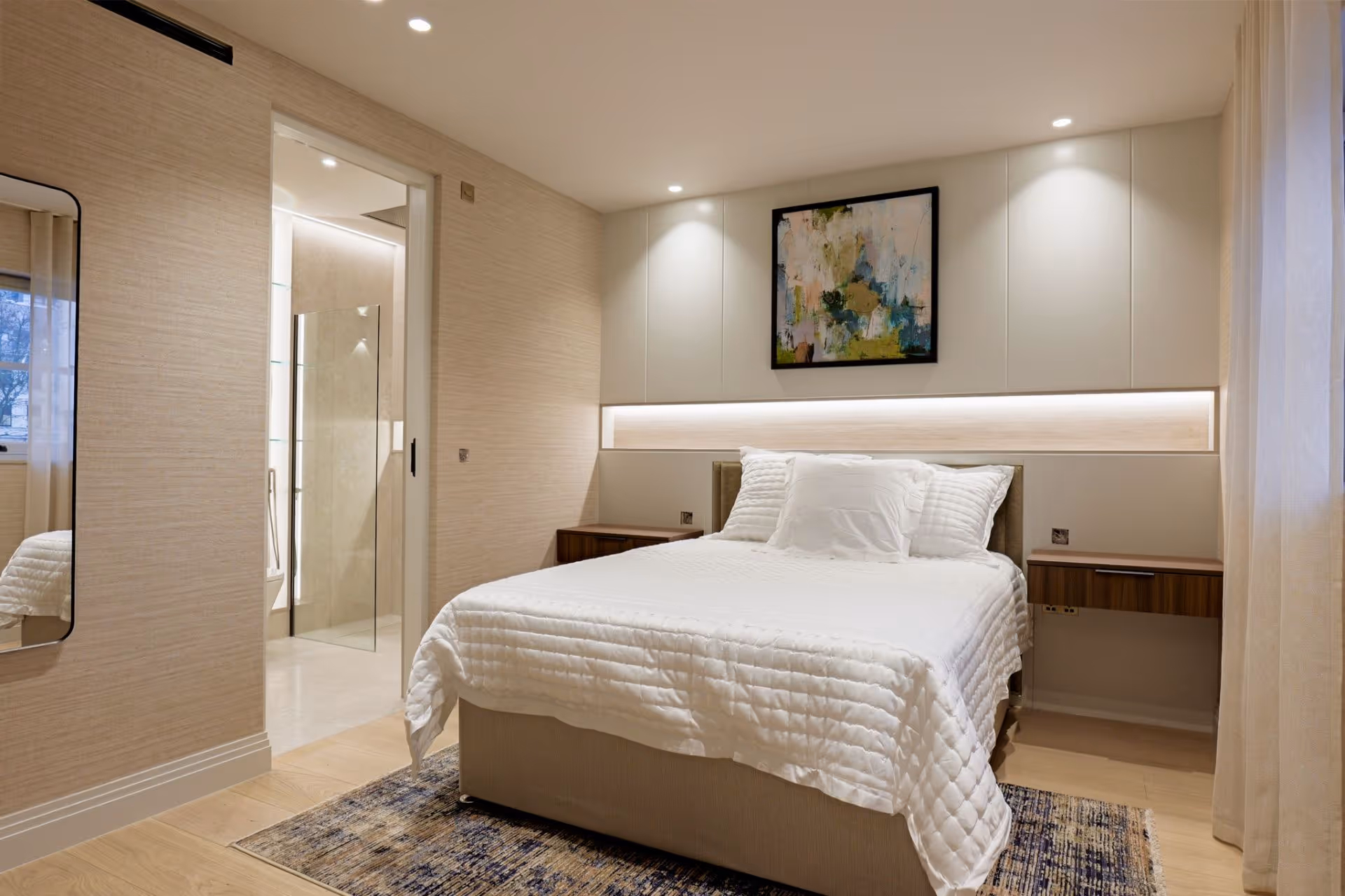 Modern bedroom with white bed, abstract art above headboard, and a doorway to an en-suite bathroom.