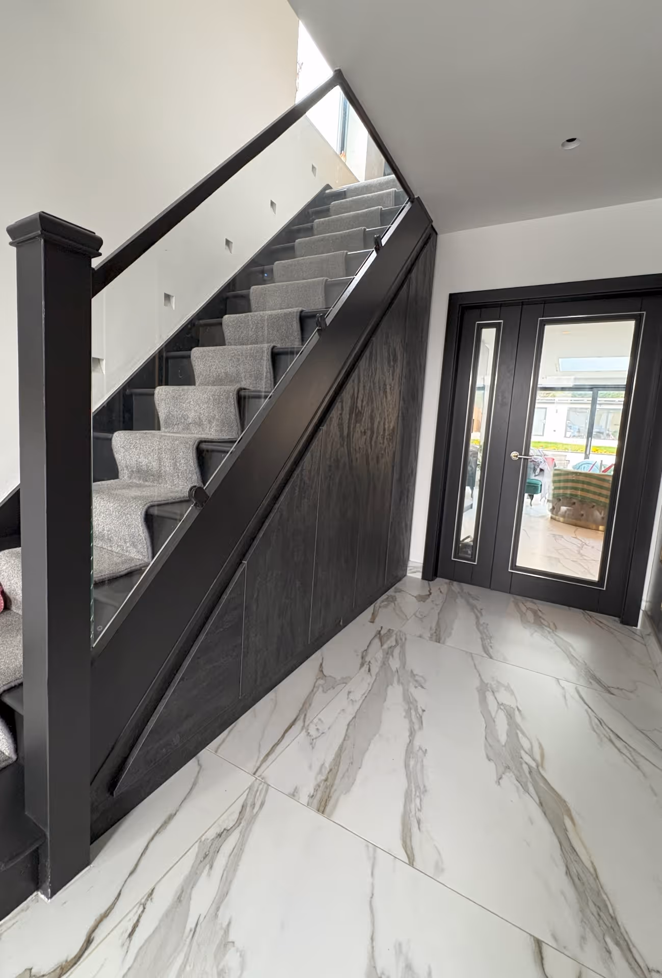 Indoor staircase with gray carpet runner, black railing and marble tile floor leading to glass-paneled double doors.