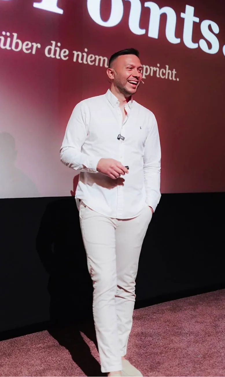 Smiling man in white shirt and pants speaking on stage with German text projected behind him.