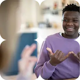 A smiling man in a purple jumper uses sign language, with a second person's hands visible in the foreground.