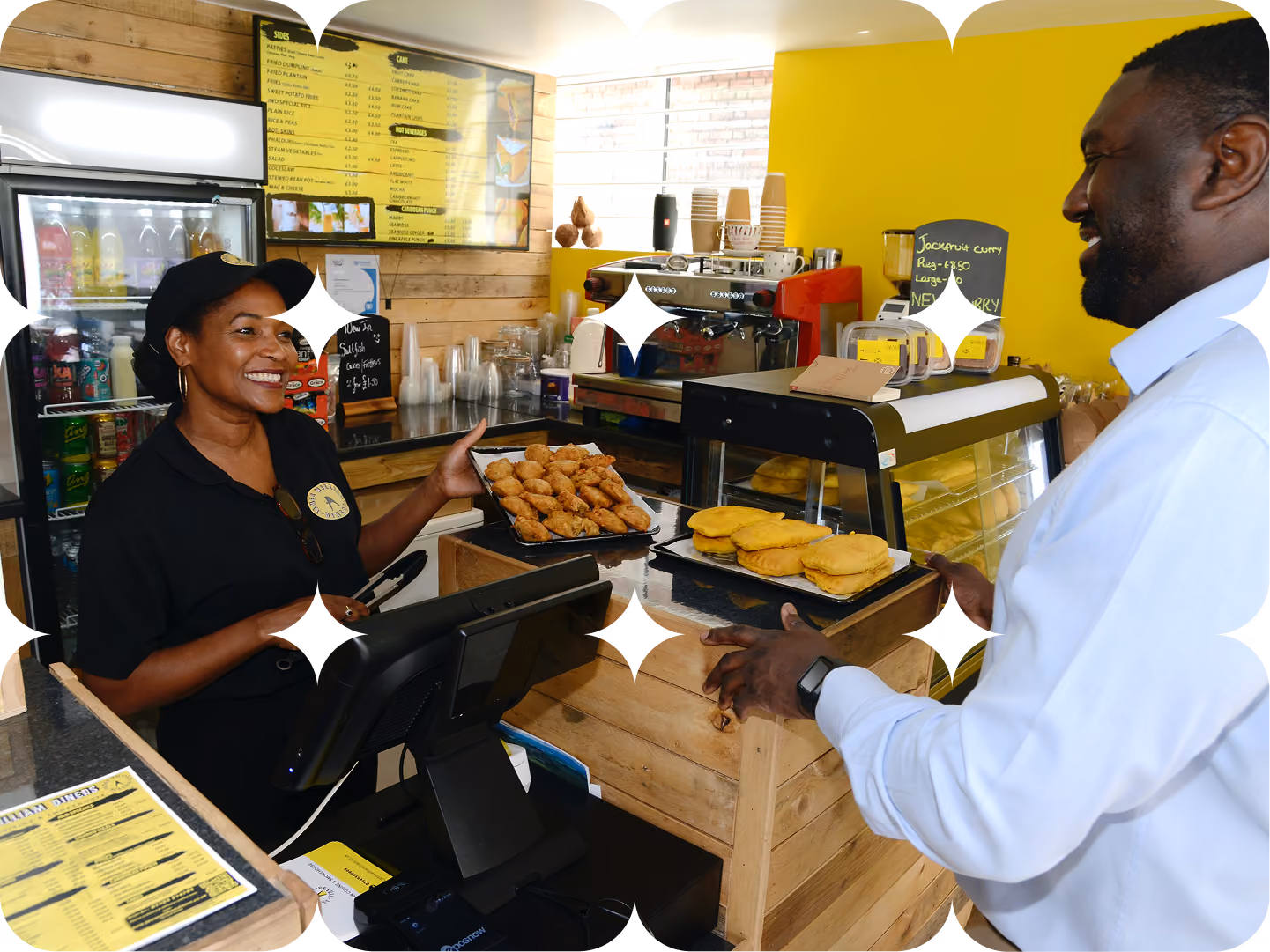 Two smiling colleagues stand behind a bakery counter, one holding a tray of pastries.