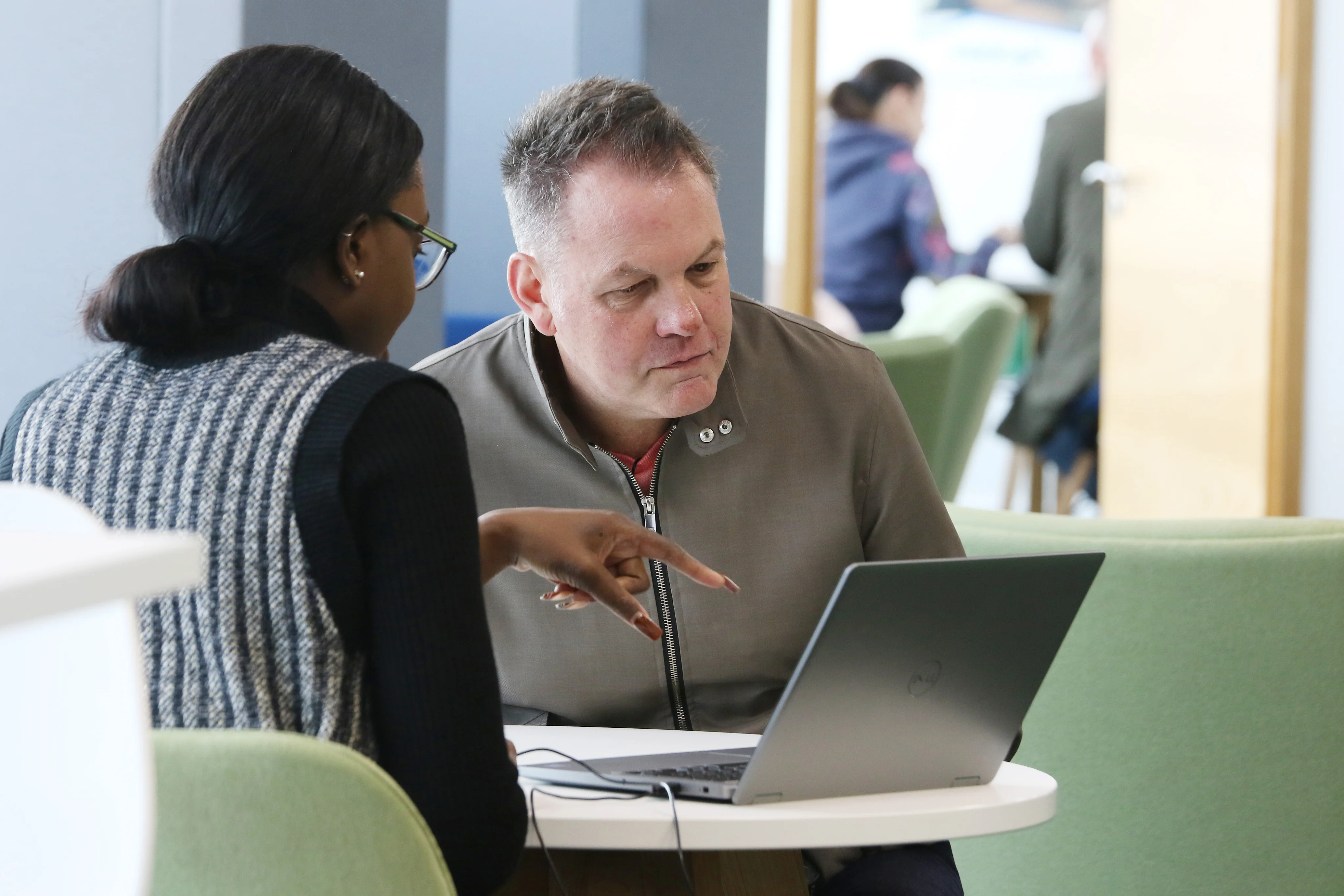 Two people sitting at a round table engaged with a laptop, one pointing at the screen.