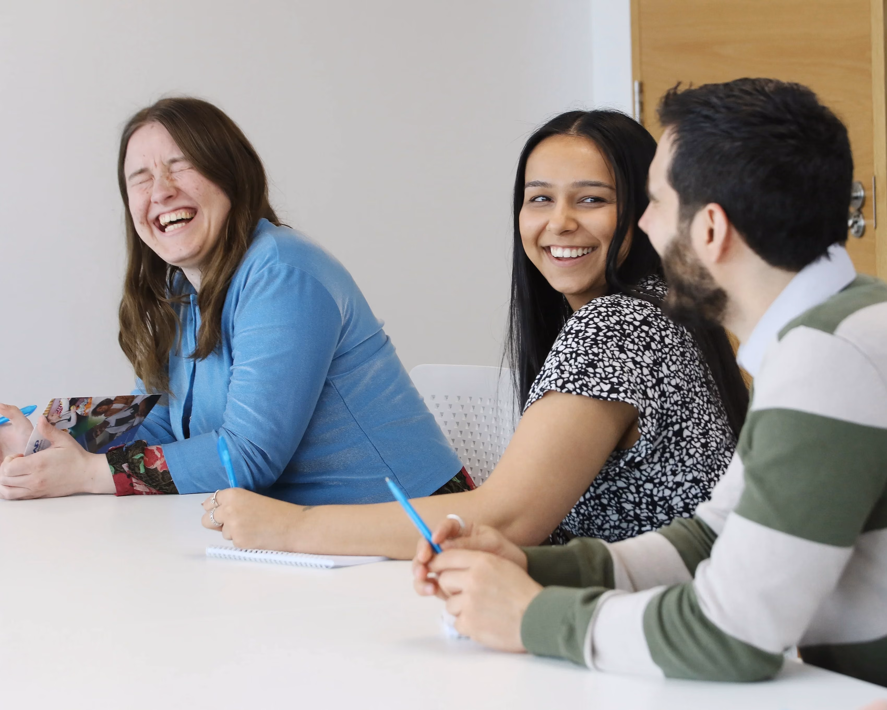 Three colleagues laugh together while talking at a table, one writing in a notebook.